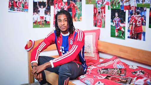 Michael Olise sitzt in einem FC Bayern-Trikot in einem Kinderbett mit Bayern-Bettwäsche in einer Nachbildung eines Thomas Müller-Kinderzimmers.