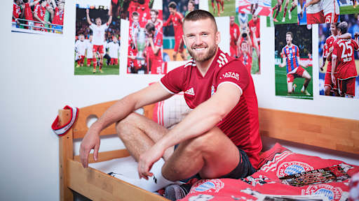 Eric Dier sitzt in einem FC Bayern-Trikot in einem Kinderbett mit Bayern-Bettwäsche in einer Nachbildung eines Thomas Müller-Kinderzimmers.