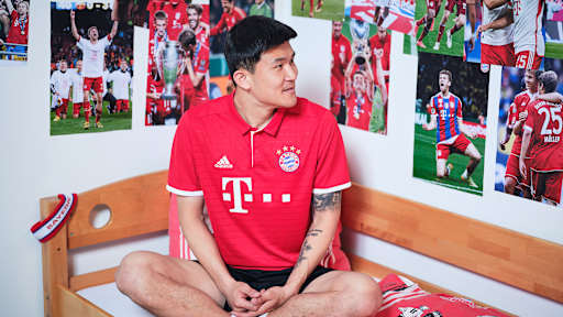Minjae Kim sitzt in einem FC Bayern-Trikot in einem Kinderbett mit Bayern-Bettwäsche in einer Nachbildung eines Thomas Müller-Kinderzimmers.