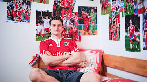 Jonas Urbig sitzt in einem FC Bayern-Trikot in einem Kinderbett mit Bayern-Bettwäsche in einer Nachbildung eines Thomas Müller-Kinderzimmers.