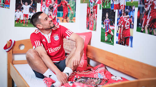 Raphael Guerreiro sitzt in einem FC Bayern-Trikot in einem Kinderbett mit Bayern-Bettwäsche in einer Nachbildung eines Thomas Müller-Kinderzimmers.