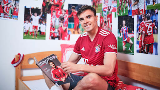 Joao Palhinha sitzt in einem FC Bayern-Trikot in einem Kinderbett mit Bayern-Bettwäsche in einer Nachbildung eines Thomas Müller-Kinderzimmers.