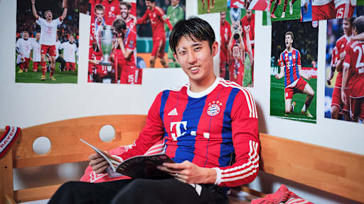 Hiroki Ito sitzt in einem FC Bayern-Trikot in einem Kinderbett mit Bayern-Bettwäsche in einer Nachbildung eines Thomas Müller-Kinderzimmers.