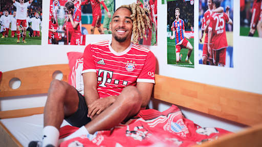 Sacha Boey sitzt in einem FC Bayern-Trikot in einem Kinderbett mit Bayern-Bettwäsche in einer Nachbildung eines Thomas Müller-Kinderzimmers.