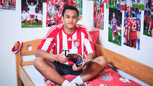 Tarek Buchmann sitzt in einem FC Bayern-Trikot in einem Kinderbett mit Bayern-Bettwäsche in einer Nachbildung eines Thomas Müller-Kinderzimmers.