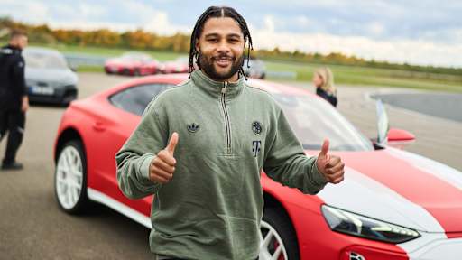 Serge Gnabry lächelt und streckt beide Dauemn nach oben vor einem roten Audi