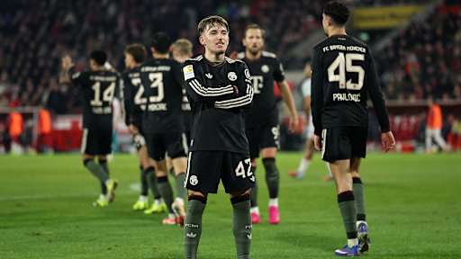 Lennart Karl steht mit verschränkten Armen vor der Kurve nach dem 3:1 im Bundesliga-Spiel des FC Bayern beim 1. FC Köln