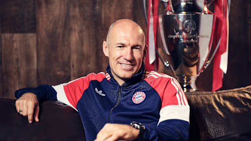 Arjen Robben grinst in einer Trainingsjacke des FC Bayern auf einer Couch.