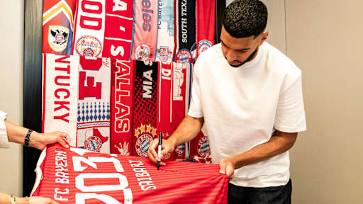 Ismael Saibari unterschreibt ein FC Bayern-Trikot mit der Zahl 2013 darauf vor aufgehängten Fan-Schals mi
