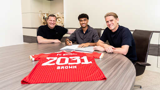 Max Eberl, Nathaniel Brown und Christoph Freund sitzen an einem Tisch, auf dem ein Vertrag und ein FCB-Trikot mit 2031 Brown liegen