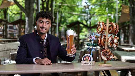 Nathaniel Brown sitzt in Tracht in einem Biergarten und hält ein Paulaner-Bier in der Hand