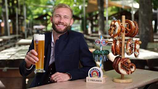 Konrad Laimer sitzt in Tracht in einem Biergarten und hält ein Paulaner-Bier in der Hand