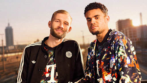 Konrad Laimer trägt das neue FC Bayern-Wiesn-Trikot 2026/27 mit farbenfrohem floralen Muster und darüber einen schwarzen Janker, daneben Luis Diaz ebenfalls im Wiesn-Trikor, im Hintergrund sind Bahngleise in München zu sehen