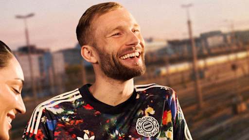 Konrad Laimer trägt lachend das neue FC Bayern-Wiesn-Trikot 2026/27 mit farbenfrohem floralen Muster, im Hintergrund sind Bahngleise in München zu sehen