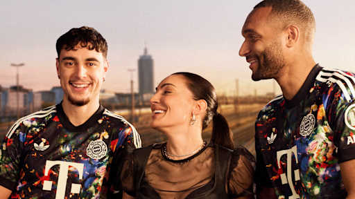 Neben einer Frau im schwarzen Dirndl tragen Jonathan Tah und Aleksandar Pavlovic das neue FC Bayern-Wiesn-Trikot 2026/27 mit farbenfrohem floralen Muster, im Hintergrund sind Bahngleise in München zu sehen