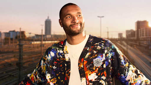 Jonathan Tah trägt die neue FC Bayern-Wiesn-Jacke 2026/27 mit farbenfrohem floralen Muster, im Hintergrund sind Bahngleise in München zu sehen