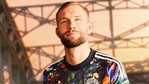 Konrad Laimer trägt das neue FC Bayern-Wiesn-Trikot 2026/27 mit farbenfrohem floralen Muster, im Hintergrund ist die Münchner Hackerbrücke zu sehen