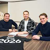 Max Eberl, Manuel Neuer und Christoph Freund sitzen an einem Tisch, Neuer unterschreibt einen Vertrag, auf dem Tisch liegt ein Trikot mit Neuer 2026
