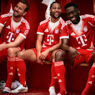 Harry Kane, Serge Gnabry und Konrad Laimer sitzen lachend in der Kabine und tragen das neue Heimtrikot des FC Bayern in Rot und Weiß mit stilisiertem M.