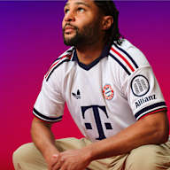 Serge Gnaby sitzt in der Hocke und trägt das neue FC Bayern Auswärtstrikot 2026/27 im Retro-Look in schlichtem Weiß mit Polo-Kragen und rot-marineblauen Details
