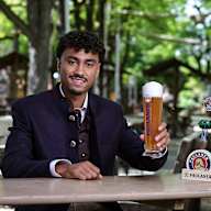 Nathaniel Brown sitzt in Tracht in einem Biergarten und hält ein Paulaner-Bier in der Hand