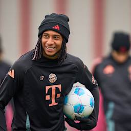 Michael Olise lächelnd mit einem Ball unter dem Arm im FC Bayern-Training vor dem Bundesliga-Spiel bei Union Berlin