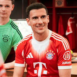 Leon Goretzka sitzt lächelnd in der Kabine und trägt das neue Heimtrikot des FC Bayern in Rot und Weiß mit stilisiertem M.