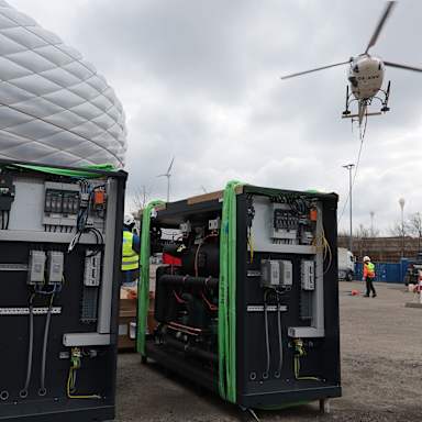 Ein Hubschrauber hebt vor der Allianz Arena ab, im Vordergrund: 2 Wärmepumpen