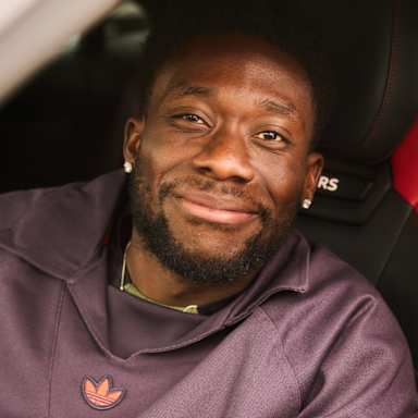 Alphonso Davies sitzt auf dem Fahrersitz seines Audi und lächelt