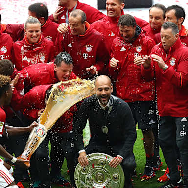 Pep Guardiola wird mit Bier überschüttet als er mit der Meisterschale und dem Trainerteam posiert.