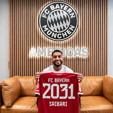 Ismael Saibari sitzt auf einem Sofa und hält lächelnd ein rotes Trikot mit der Aufschrift FC Bayern 2031 Saibari hoch