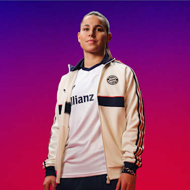 Lena Oberdorf trägt das neue FC Bayern Auswärtstrikot 2026/27 im Retro-Look in schlichtem Weiß mit Polo-Kragen und rot-marineblauen Details, darüber eine weiße Sportjacke des FC Bayern