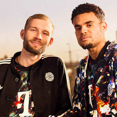 Konrad Laimer trägt das neue FC Bayern-Wiesn-Trikot 2026/27 mit farbenfrohem floralen Muster und darüber einen schwarzen Janker, daneben Luis Diaz ebenfalls im Wiesn-Trikor, im Hintergrund sind Bahngleise in München zu sehen