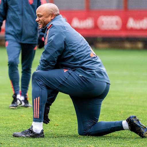 Vincent Kompany kniet im Training des FC Bayern auff dem Rasen und lacht.