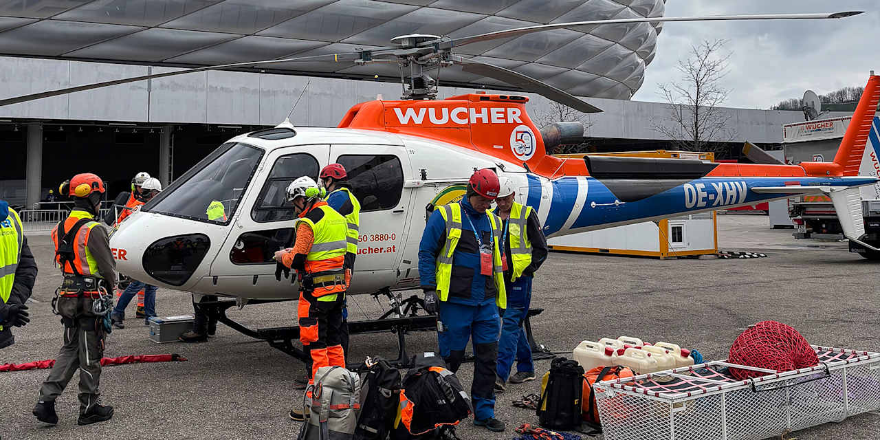 Arbeiter stehen vor einem Hubschrauber, im Hintergrund die Allianz Arena