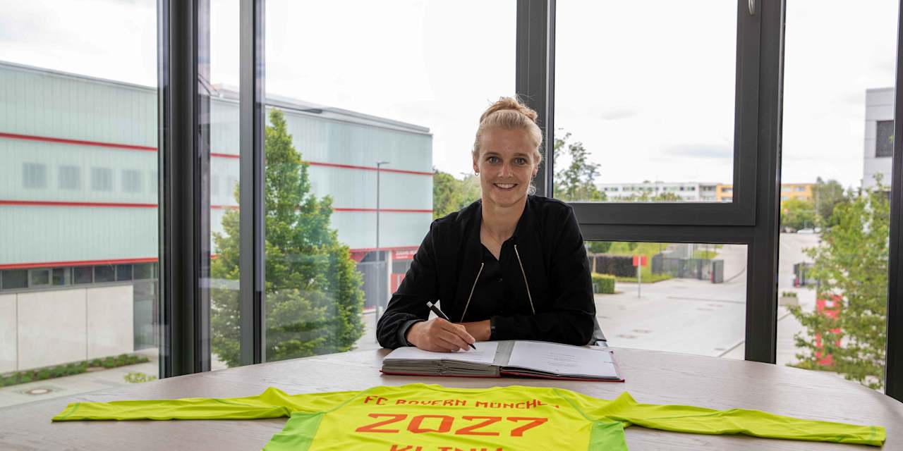 Anna Klink sits at a table and signs her contract. She smiles at the camera, in front of her a shirt with the contract term 2027.