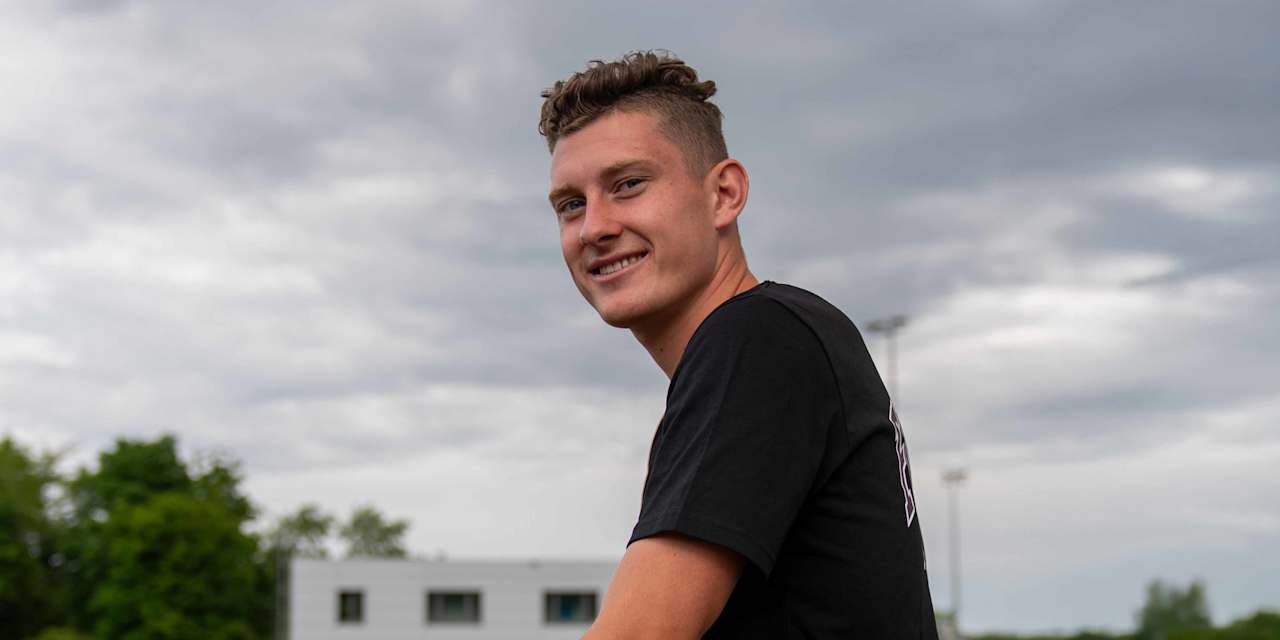Alt-Tag: David Heindl steht mit verschränkten Armen und einem Lächeln vor dem FC Bayern Logo