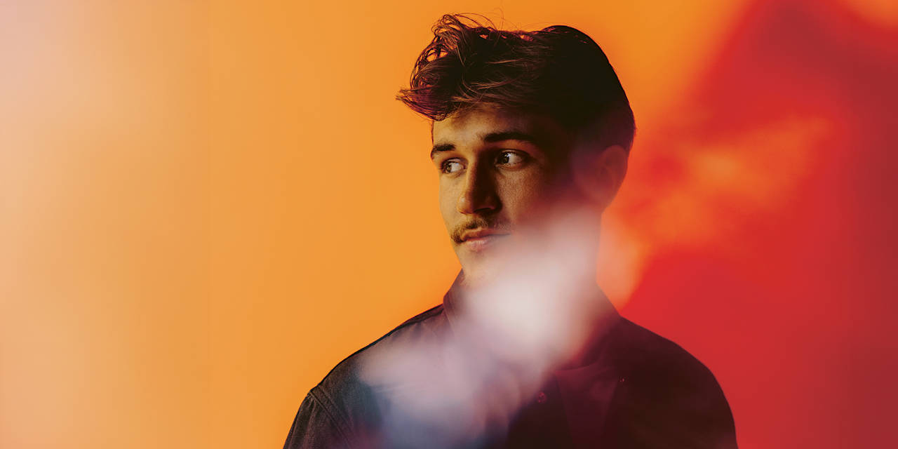 Tom Bischof looks to the side in front of a wall illuminated in yellow and red.