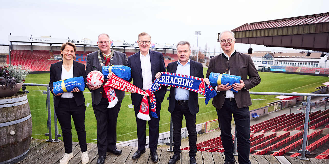 Bianca Rech, Wolfgang Panzer, Jan-Christian Dreesen, Manfred Schwabl und Jürgen Muth halten zwei zusammengeknotete Schals des FC Bayern und der SpVgg Unterhaching im Sportpark Unterhaching.