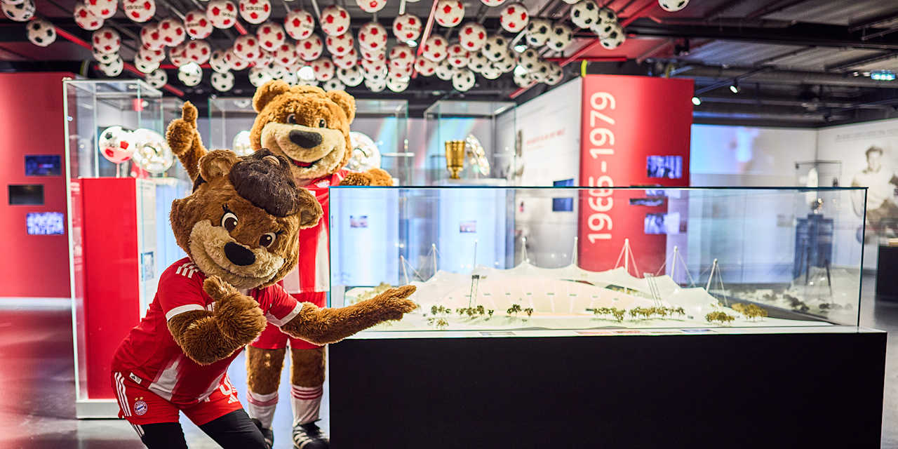 Die Maskottchen Berni und Mia stehen vor einem Ausstellungskasten im FC Bayern Museum.