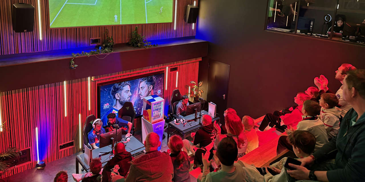 In einer Veranstaltungshalle spielen zwei E-Sportler gegeneinander Fifa, der Screen wird groß auf eine Leinwand übertragen für die Fans.