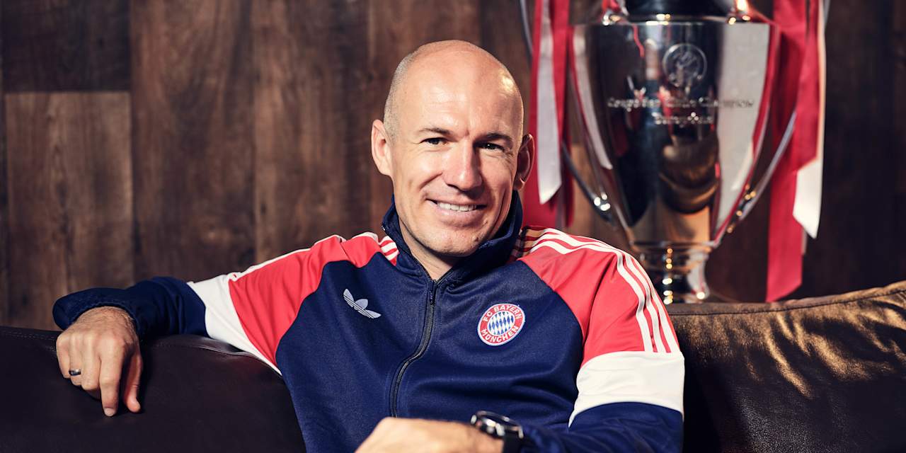 Arjen Robben grinst in einer Trainingsjacke des FC Bayern auf einer Couch.