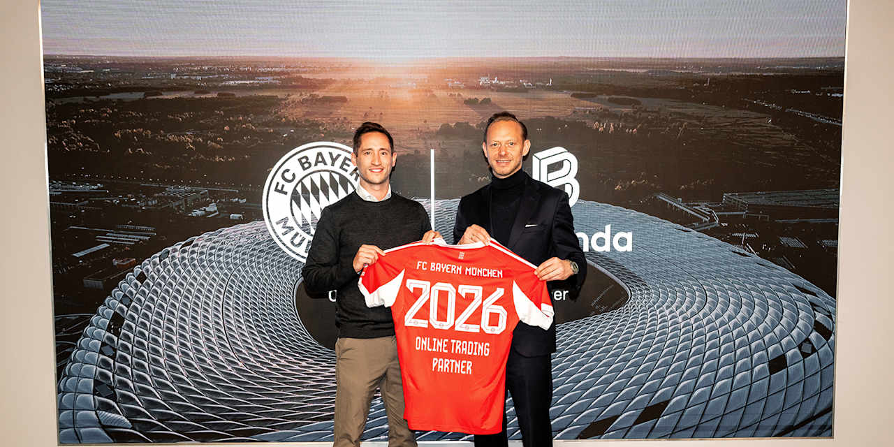 Dominik Beier, CCO Bitpanda, und Rouven Kasper, Vorstand Marketing & Vertrieb des FC Bayern, halten ein Rotes Trikot mit der Aufschrift 2026 Online Trading partner hoch