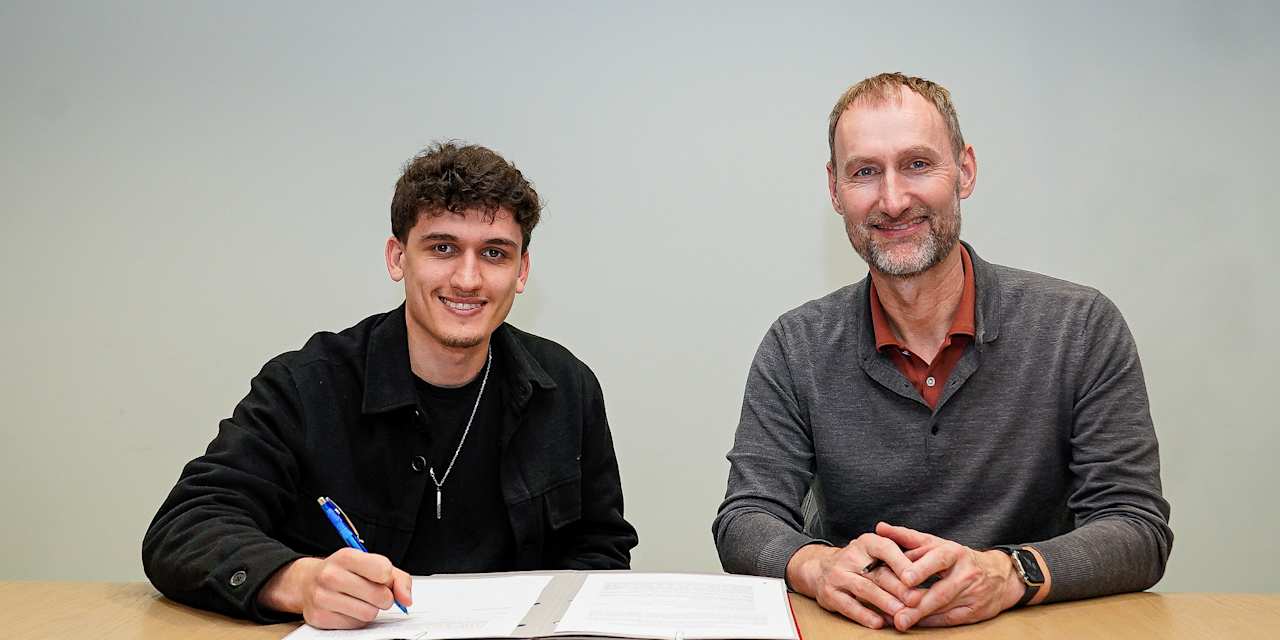 Younes Aitamer unterzeichnet seinen Vertrag und lächelt mit Jochen Sauer in die Kamera.