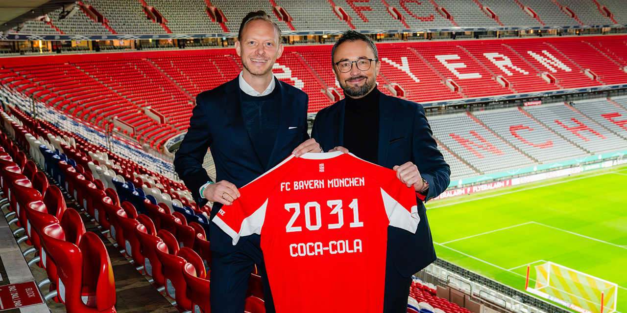 Rouven Kasper und Andreas Laub halten in der Allianz Arena ein FC Bayern-Trikot mit dem Aufdruck 2031 Coca-Cola hoch