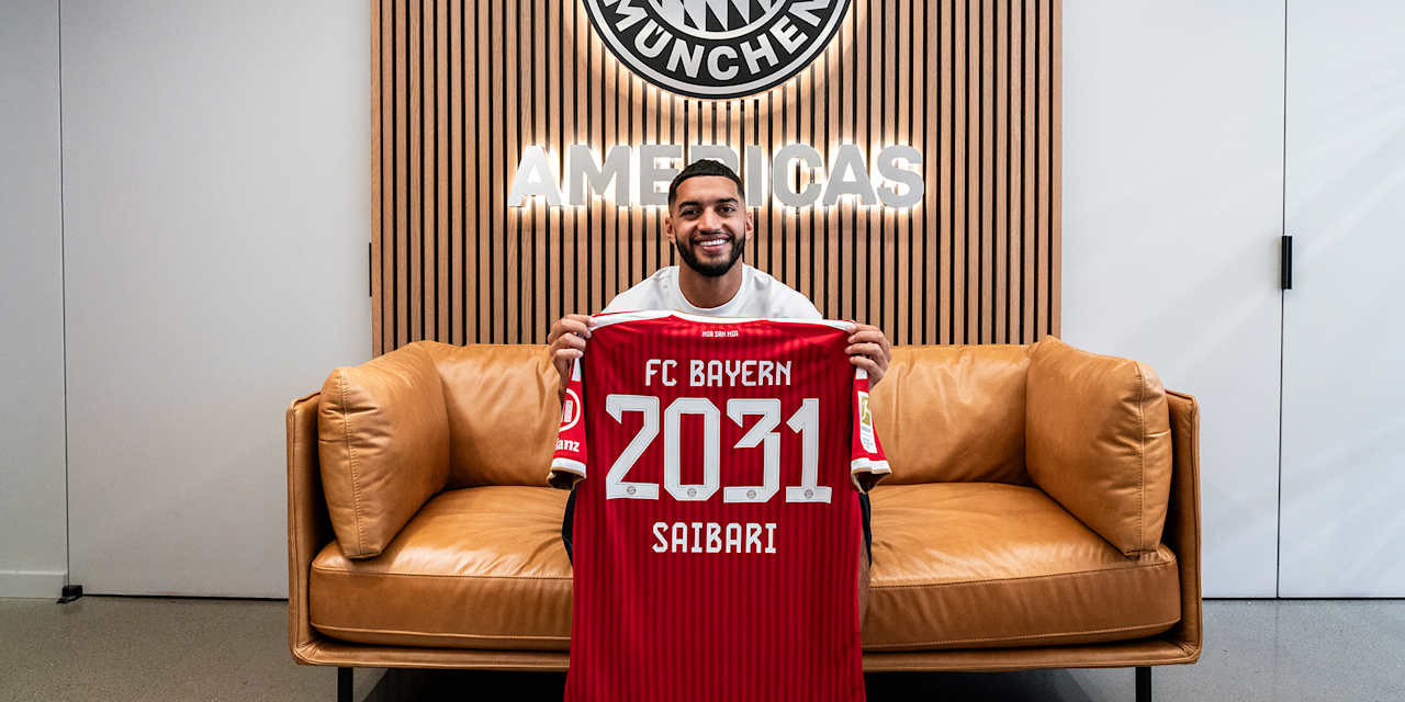 Ismael Saibari sitzt auf einem Sofa und hält lächelnd ein rotes Trikot mit der Aufschrift FC Bayern 2031 Saibari hoch