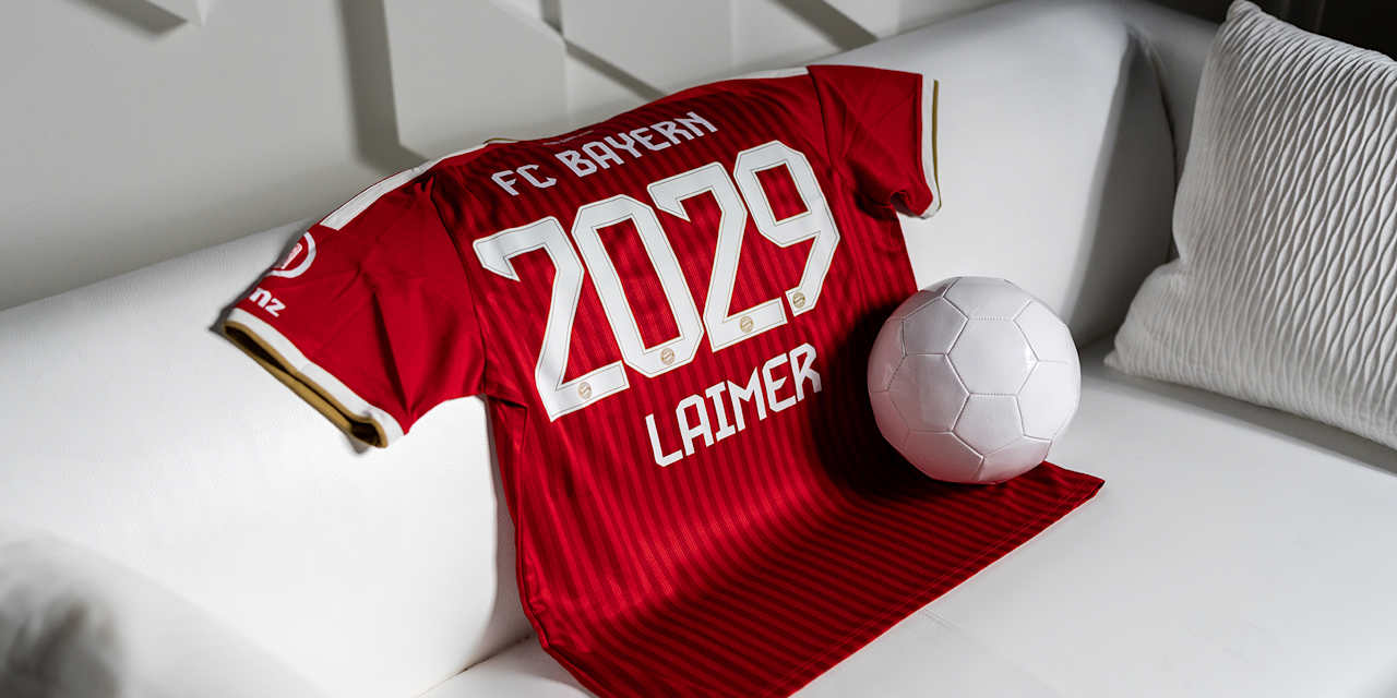 Ein rotes Trikot mit der Aufschrift FC Bayern Laimer 2029 liegt auf einer weißen Couch in einem weißen Raum neben einem weißen Ball