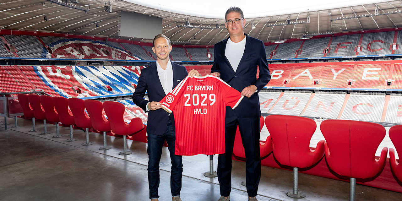 Rouven Kasper und Christian Krensel halten in der Allianz Arena ein rotes Trikot mit der Aufschrift FC Bayern 2029 HYLO hoch