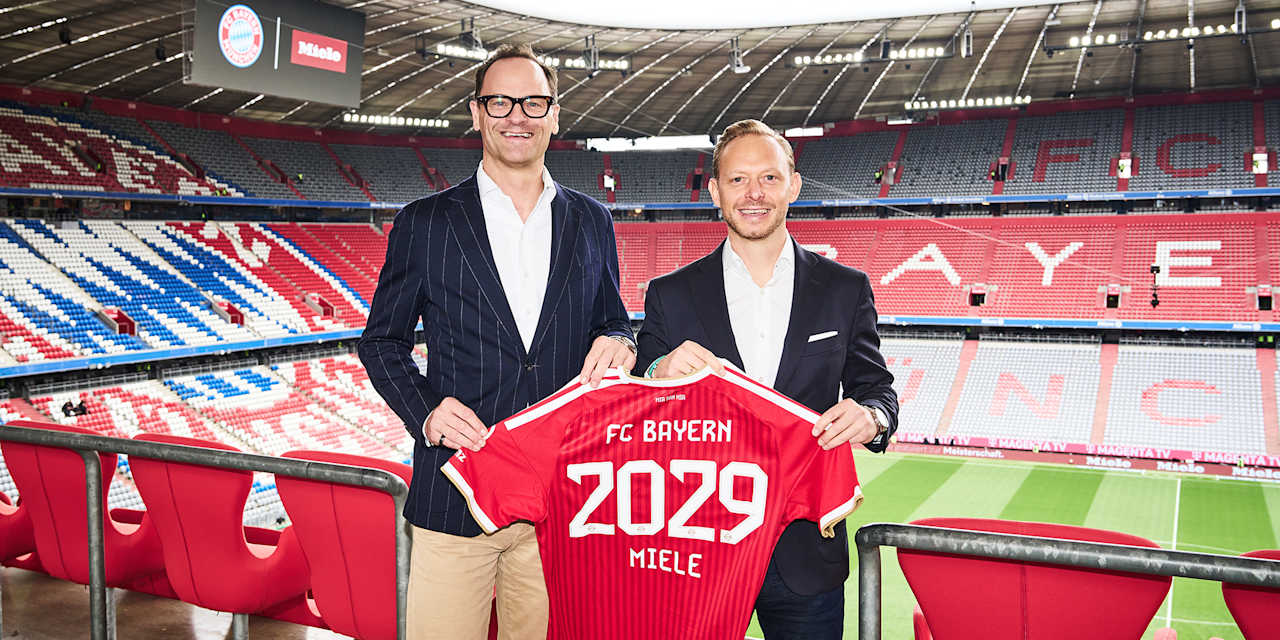Rouven Kasper und Dr. Axel Kniehl halten ein FC Bayern Trikot mit 2029 Miele in der Allianz Arena