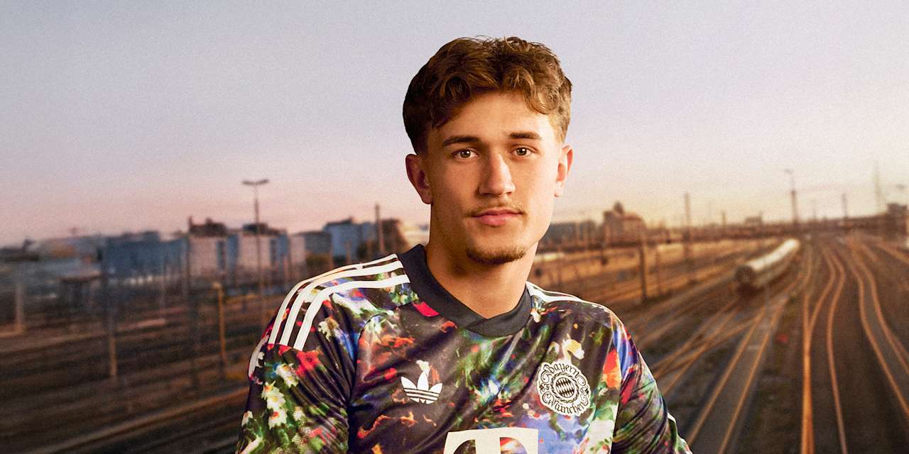 Tom Bischof trägt das neue FC Bayern-Wiesn-Trikot 2026/27 mit farbenfrohem floralen Muster, im Hintergrund sind Bahngleise in München zu sehen